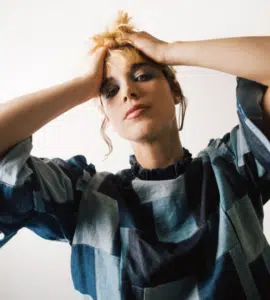 A person wearing a patchwork denim top holds their hands on their head and looks toward the camera against a plain white background.